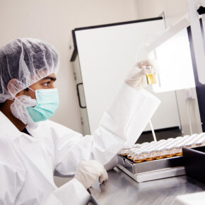 Biologics CMO technician sitting in a lab with a facemask, hair and labcoat visually inspecting vials of drug product