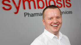 Headshot of John McCormick, member of the Symbiosis leadership team with Symbiosis branded signage behind him