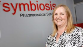 Headshot of Lorraine Den-Kaat with Symbiosis Pharmaceutical Services signage in the background