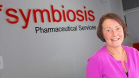 Headshot of Alison Clayton wearing a pink cardigan on front of a Symbiosis Pharmaceuticals wall sign