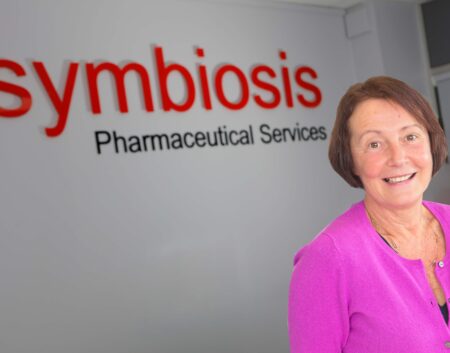 Headshot of Alison Clayton wearing a pink cardigan on front of a Symbiosis Pharmaceuticals wall sign