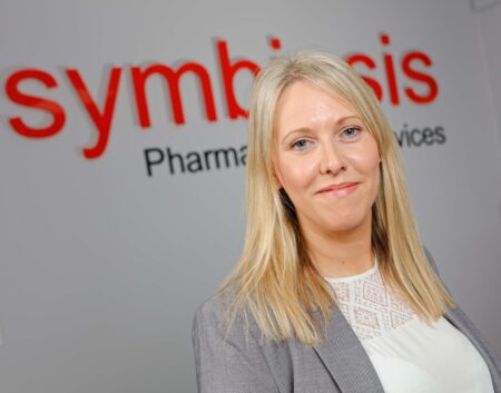 Member of the Symbiosis leadership team, Cheryl Ballentyne standing on front of Symbiosis signage wearing a grey suit