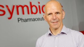 Headshot of Colin MacKay, member of the Symbiosis leadership team wearing a blue shirt standing on front of Symbiosis signage