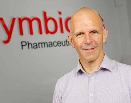 Headshot of Colin MacKay, member of the Symbiosis leadership team wearing a blue shirt standing on front of Symbiosis signage